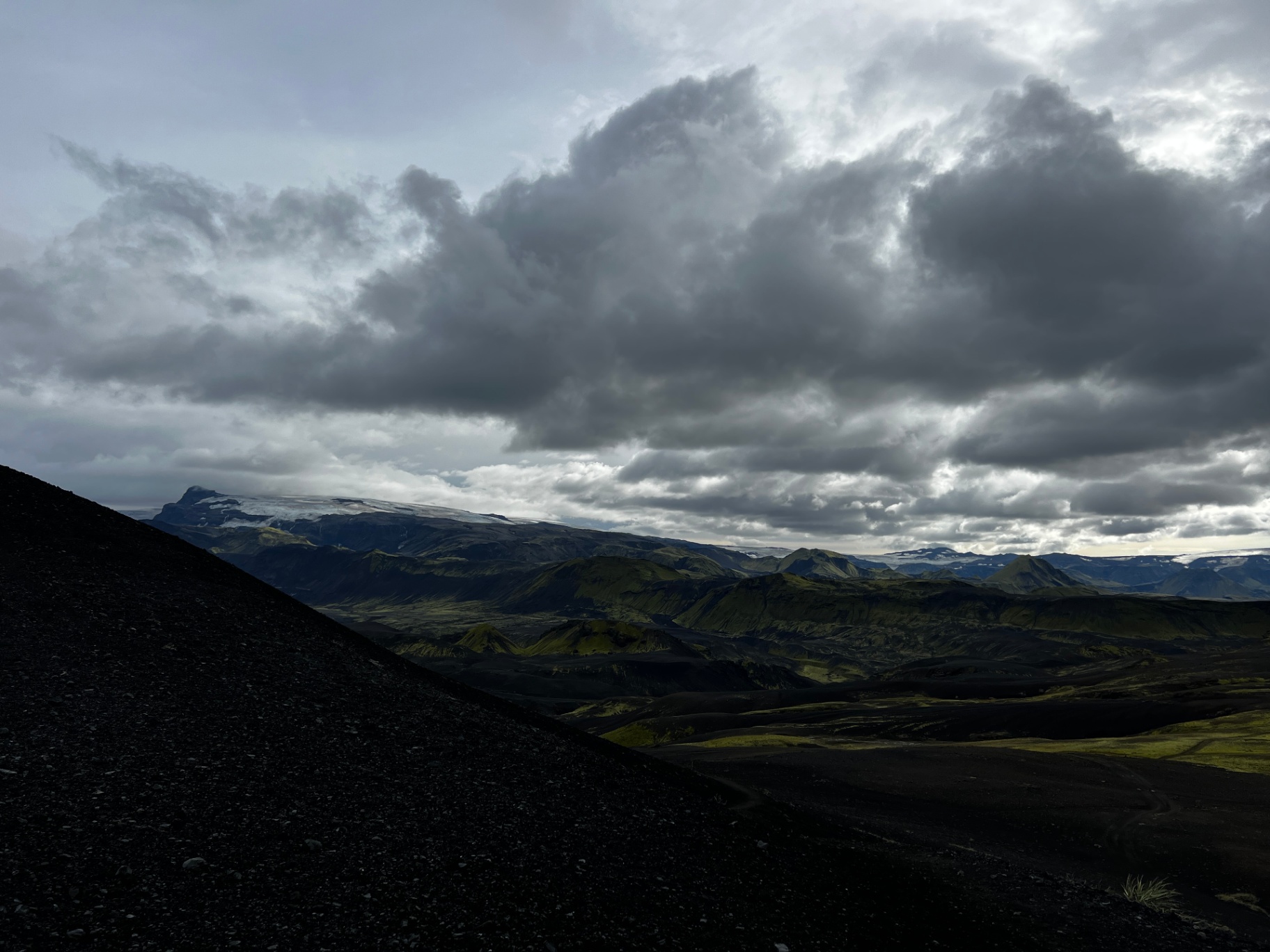 A dark volcanic slope descends toward a distant glacier under a heavy dusk sky; the day ending in the mountains.