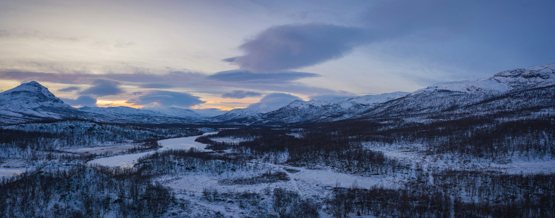 A wide snow-covered valley at dusk, a frozen river winding between low mountains under lenticular clouds: the long view, the patient season.